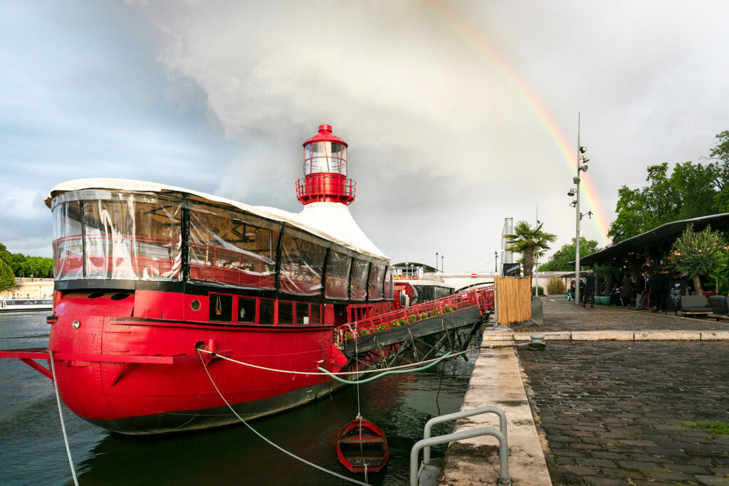 Le Bateau Phare moored at the quay, with its red lighthouse and a rainbow in the sky